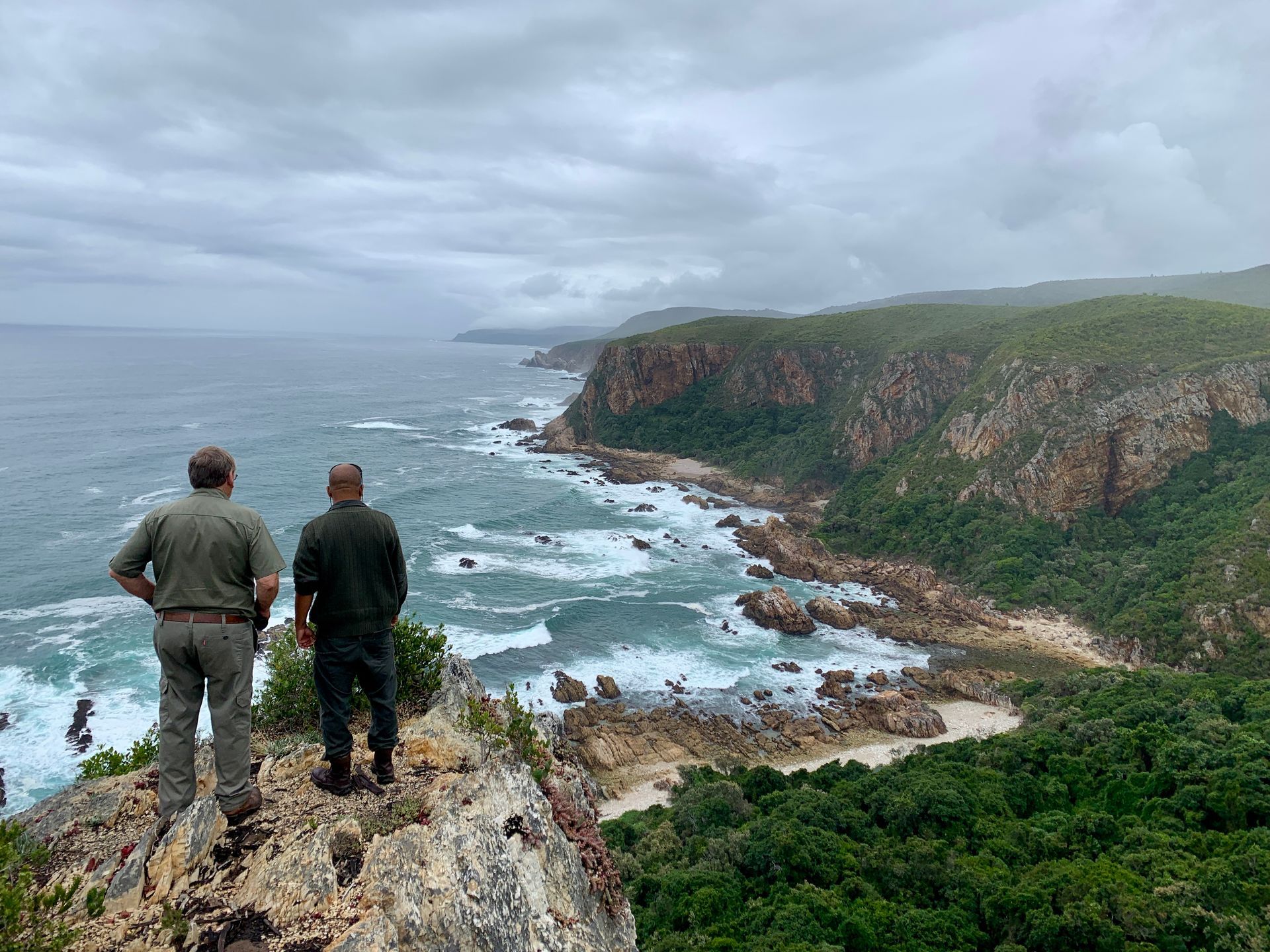 Two people overlooking a rocky coastline with crashing waves and a cloudy sky.