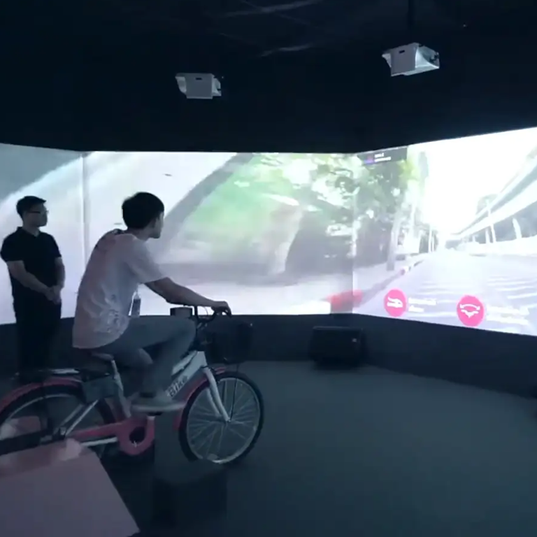 Person riding a pink bike in an indoor VR cycling simulator with two onlookers and a curved screen