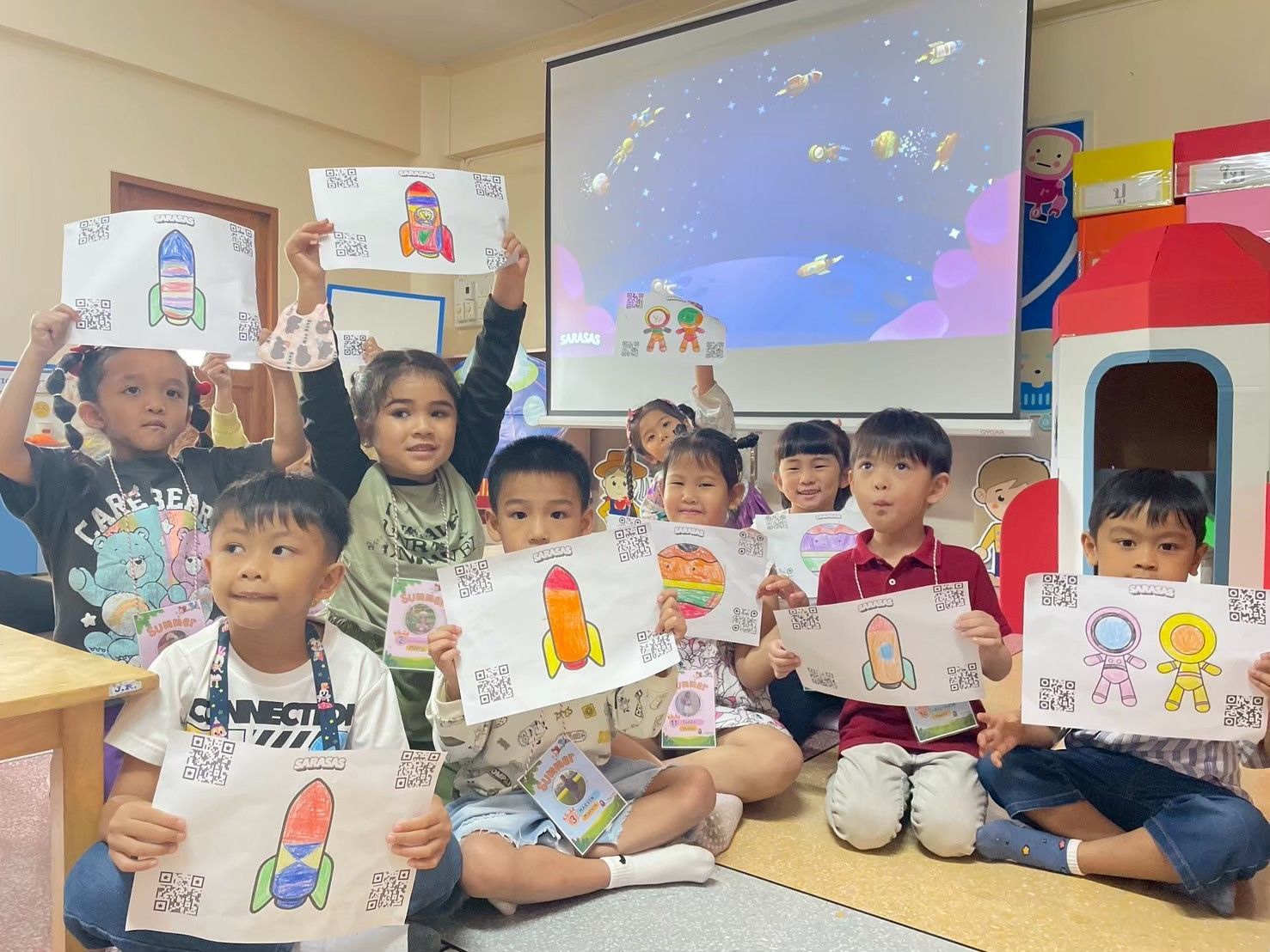 Children holding drawings in a classroom, seated in front of a projector screen and colorful play structures.