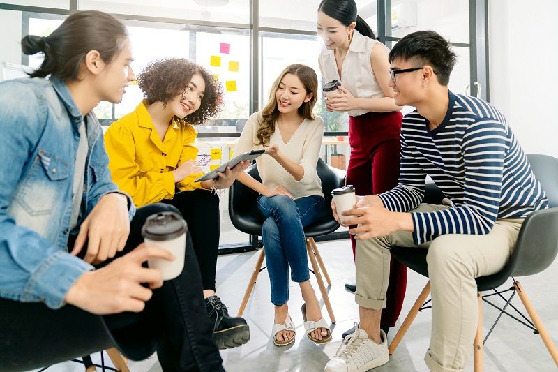 Five coworkers chatting and smiling in a bright office lounge, holding coffee cups and a tablet