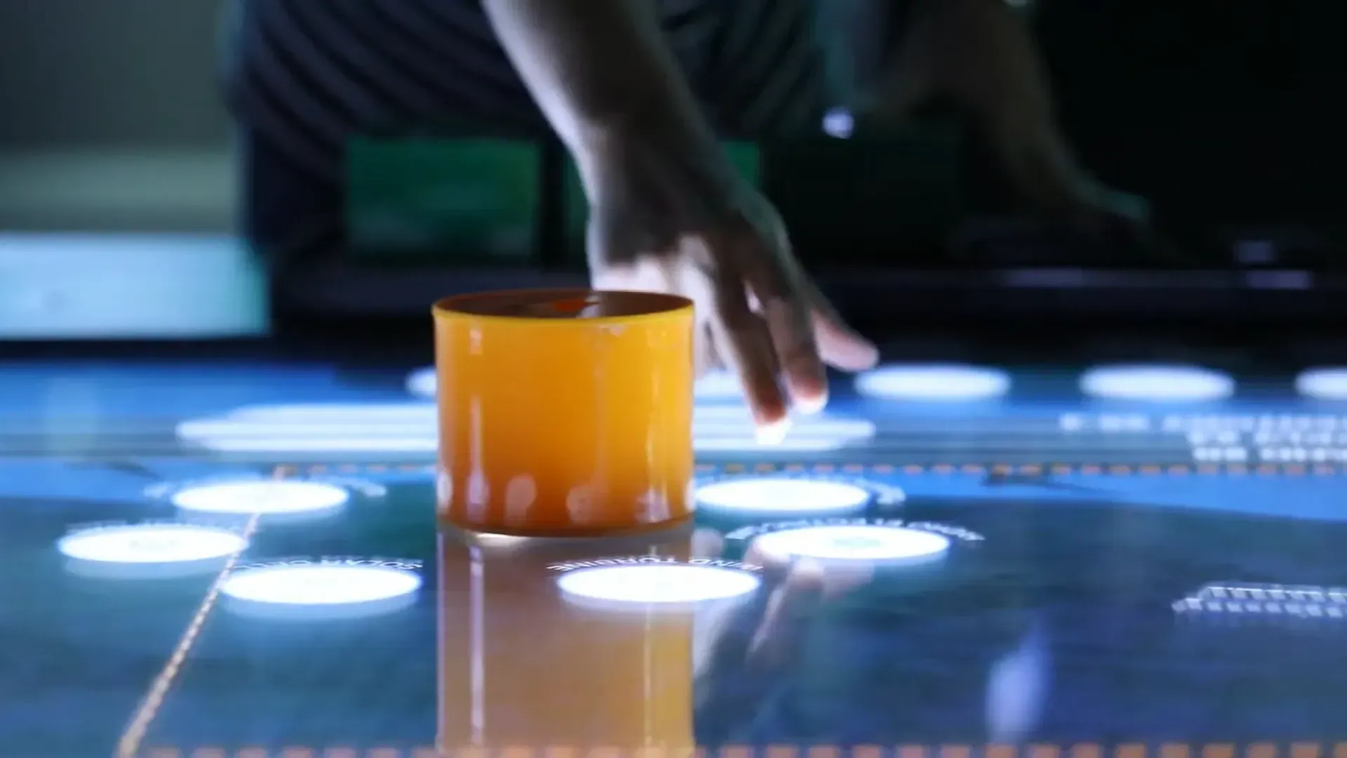 Hand reaching toward an orange cup on a glowing touchscreen table with blue-white circles