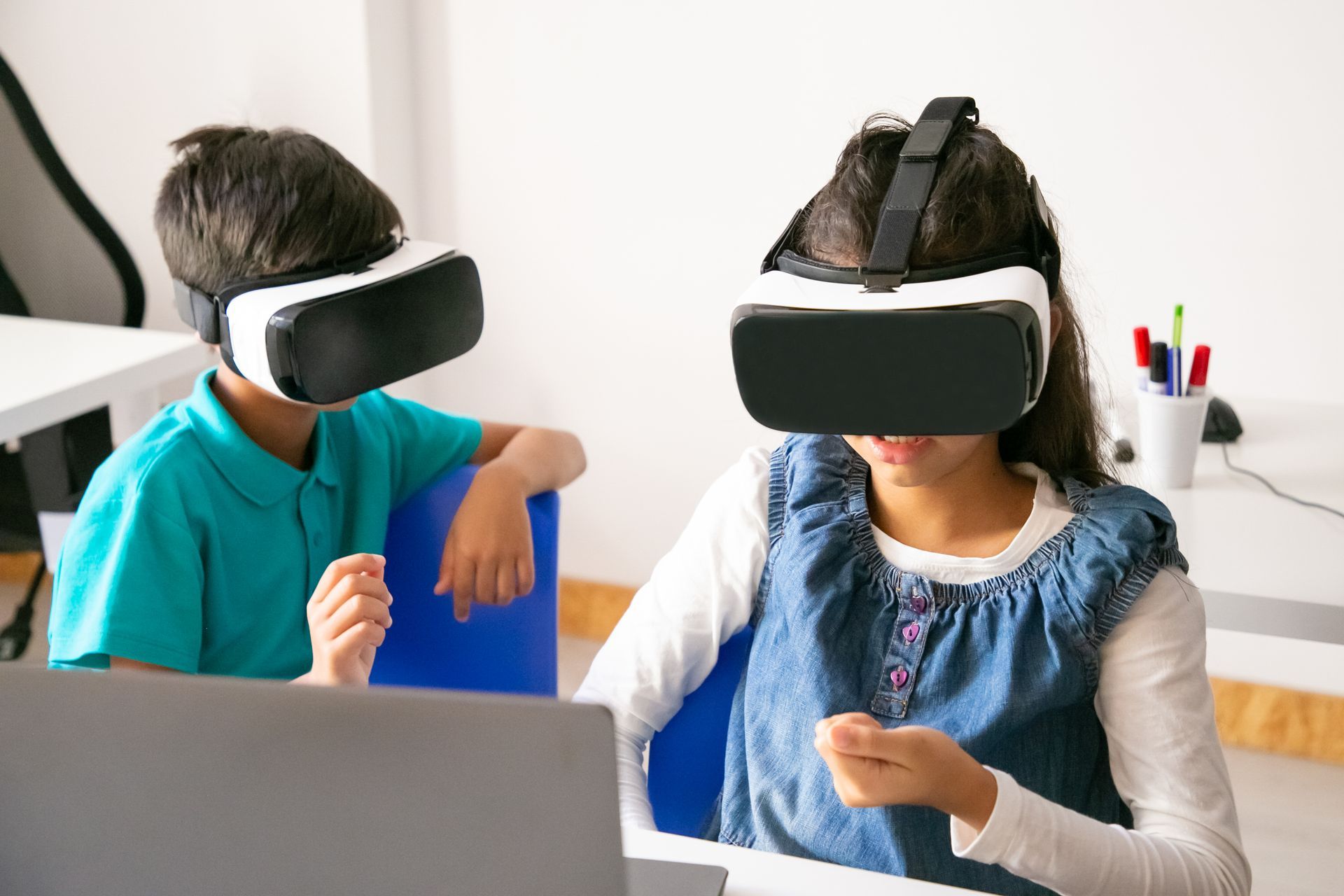 Two people wearing VR headsets sit at a table, gesturing while using a laptop in a bright room.