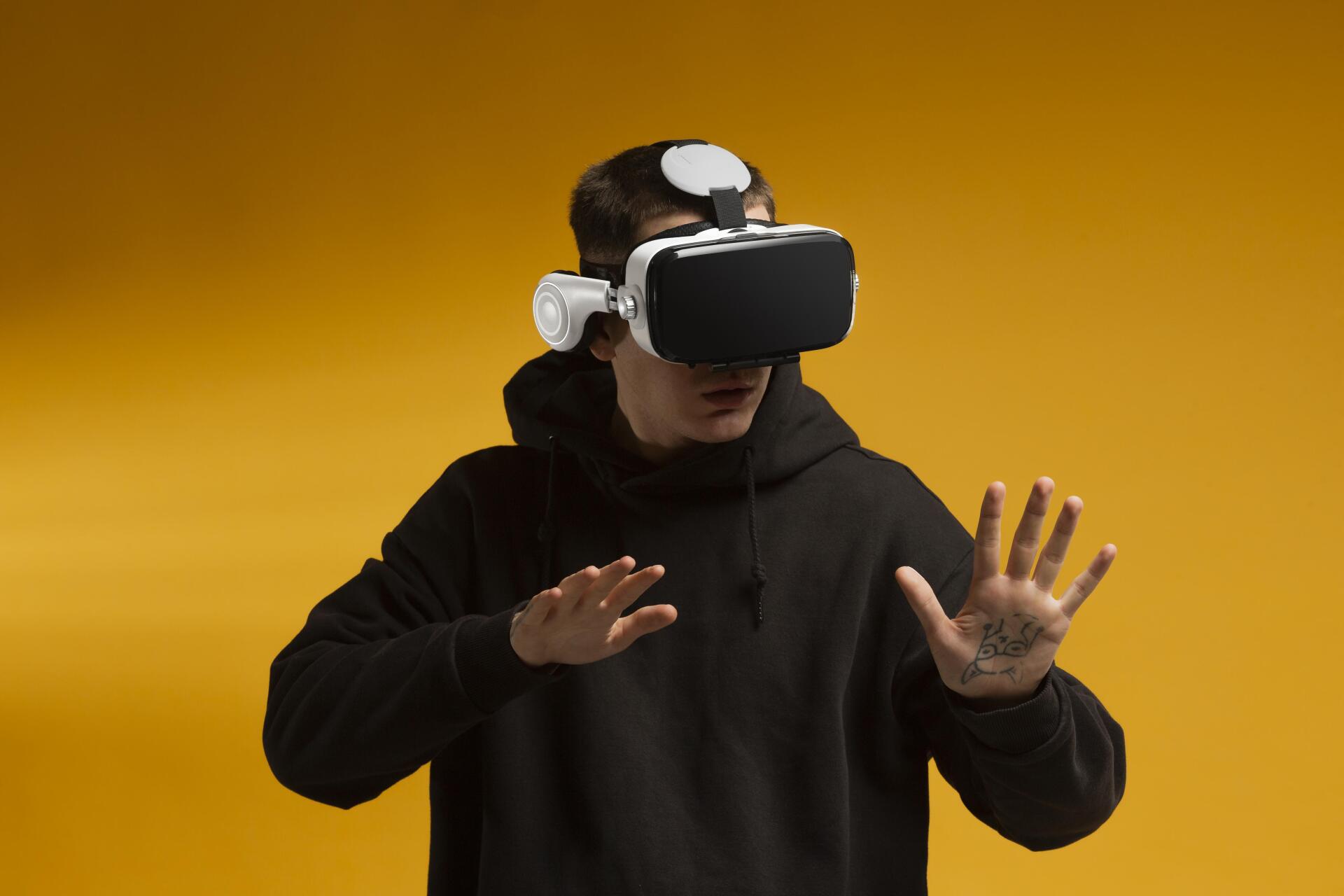 Person wearing a VR headset and black hoodie, reaching out with both hands against a yellow background