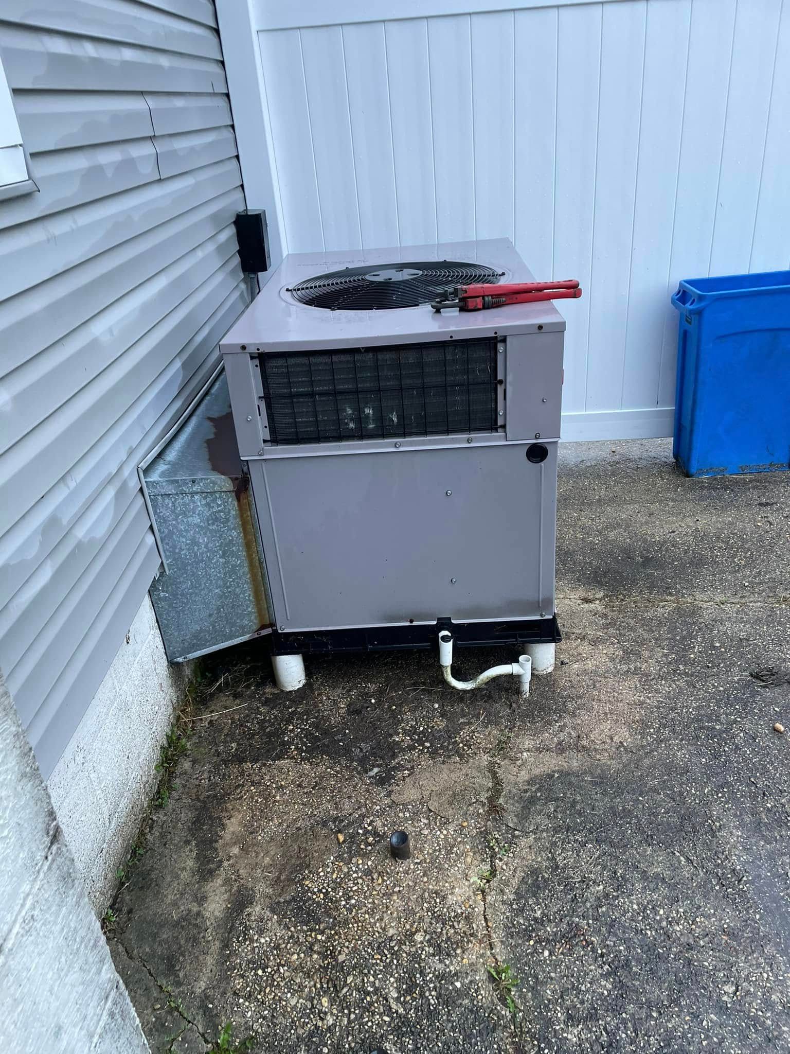Outdoor HVAC unit beside a house wall, with tools on top and a blue bin in the background.