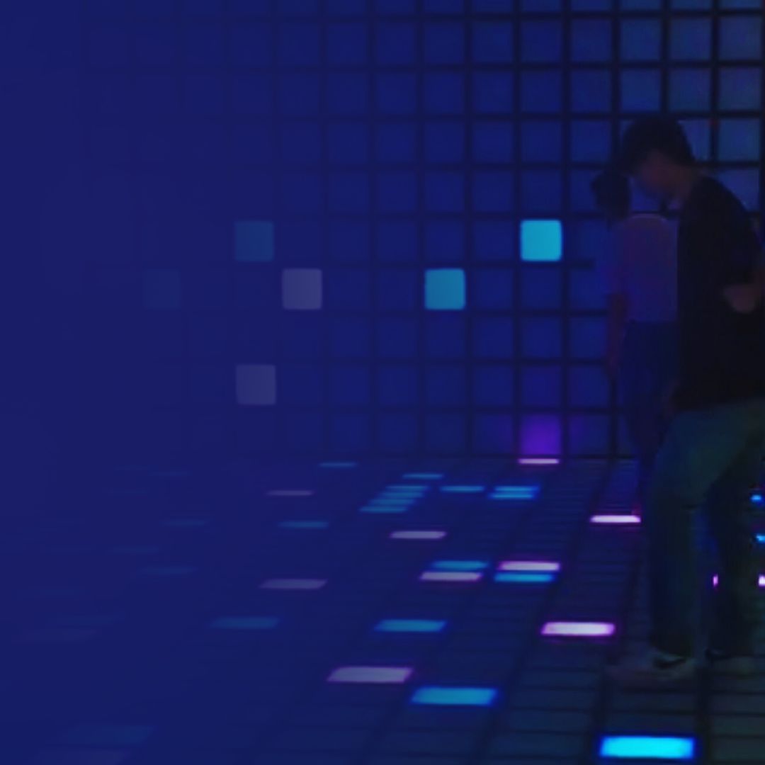 A person walks across an illuminated floor with glowing blue and purple squares against a dark, grid-patterned wall.
