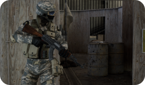 A soldier in camouflage gear holds an assault rifle while standing in a wooden training facility near two metal barrels.