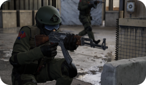 A soldier in green tactical gear kneels, aiming a rifle, with another soldier moving in the background of a gray facility.