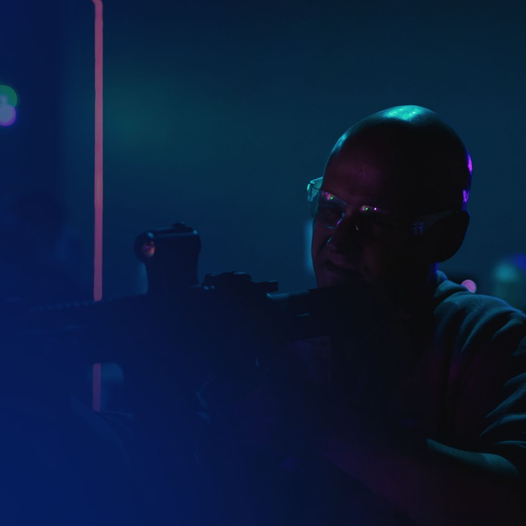 A bald person wearing safety glasses aims a rifle in a dark, blue-lit setting.