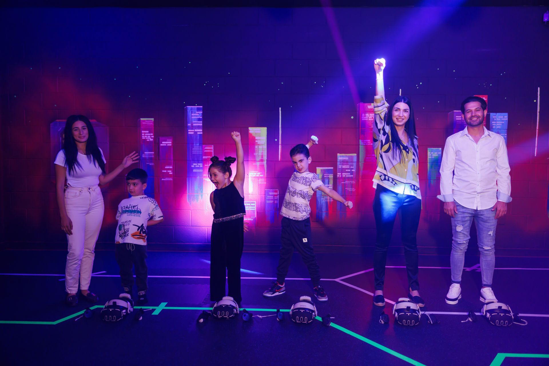A group of six people stand on a stage with neon floor lighting, some posing with arms raised under blue and purple lights.
