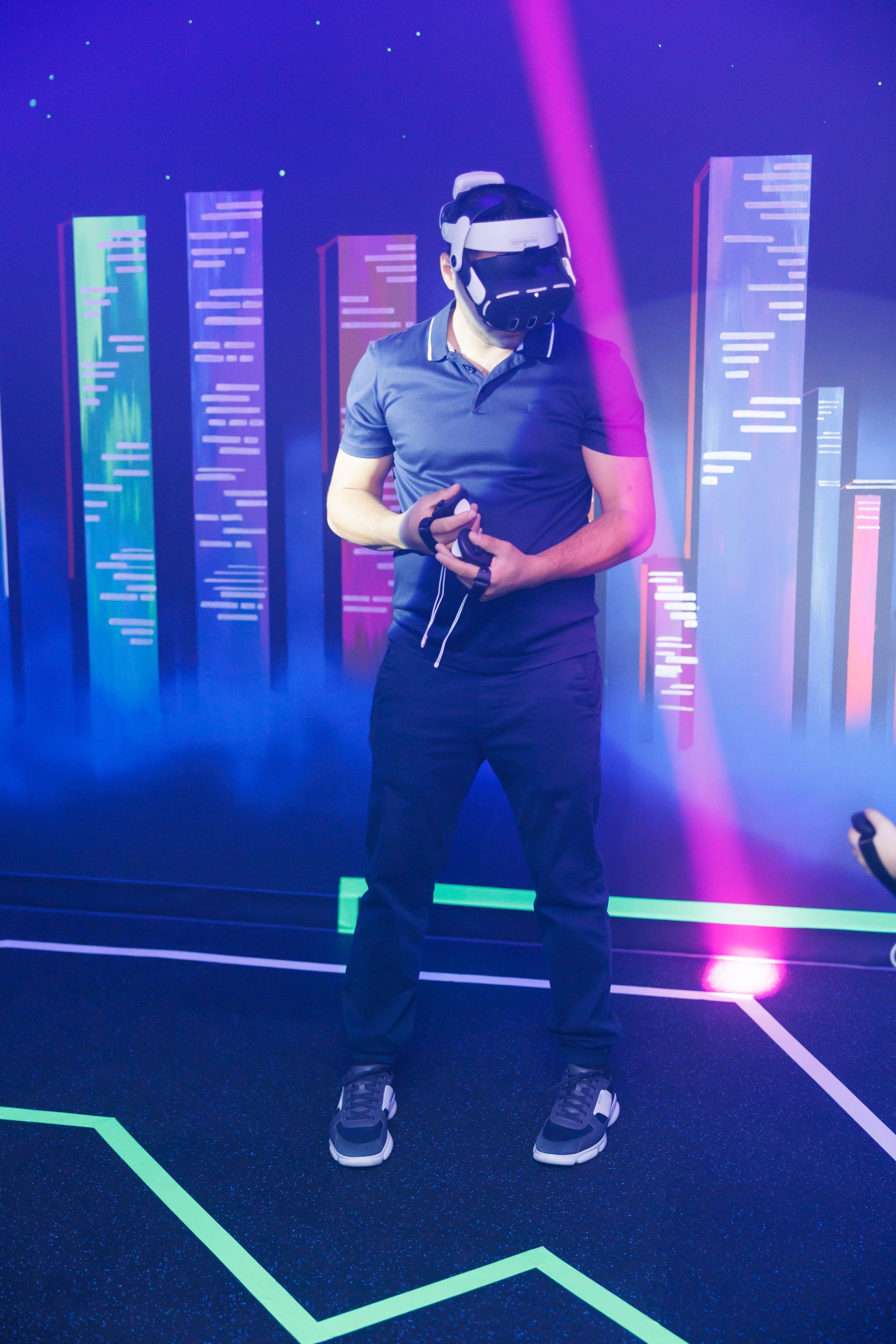 A person in dark clothing stands in a neon-lit, futuristic space wearing a VR headset and holding controllers.