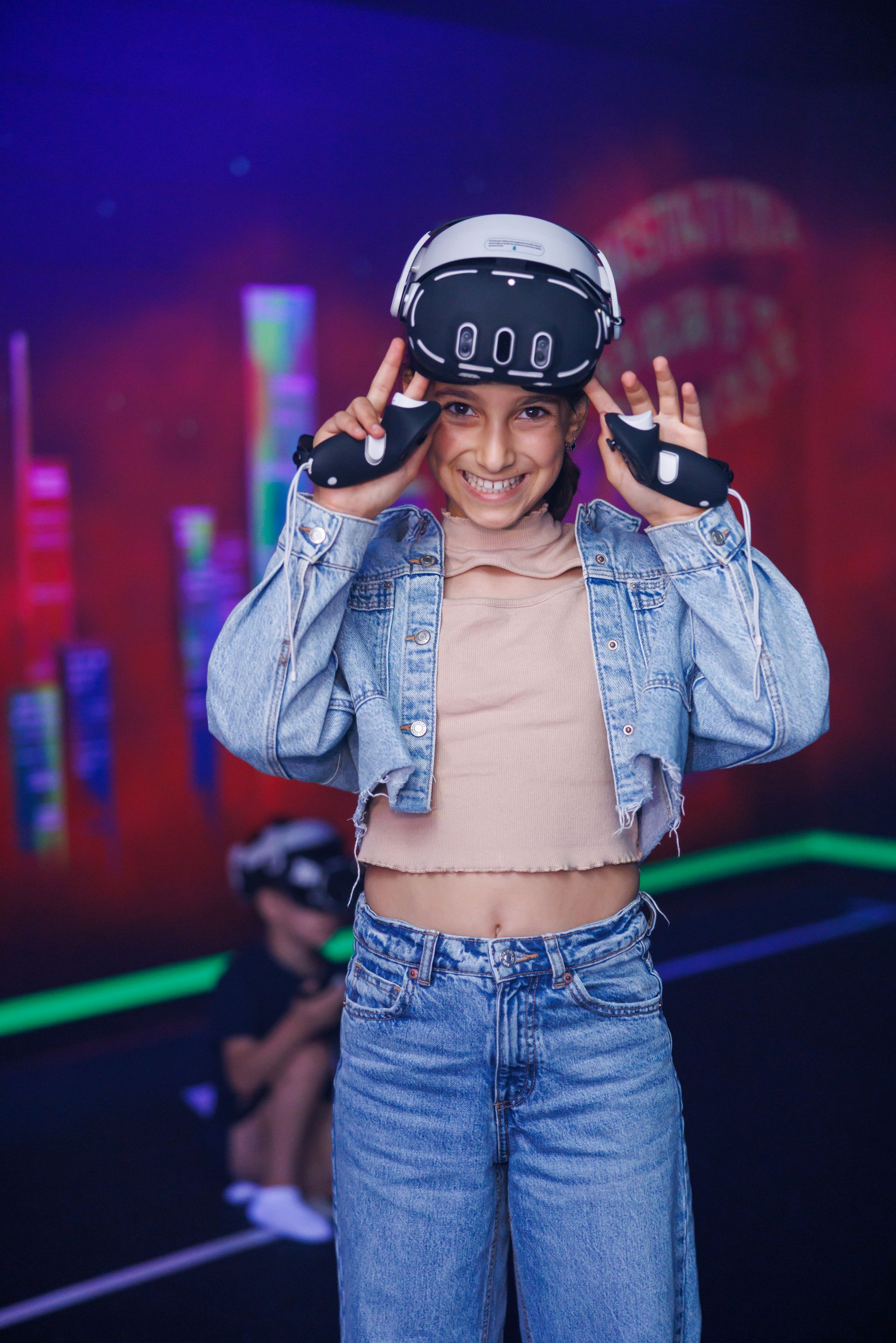 A smiling person wearing a VR headset and holding controllers, dressed in a denim jacket, standing in a gaming arena.