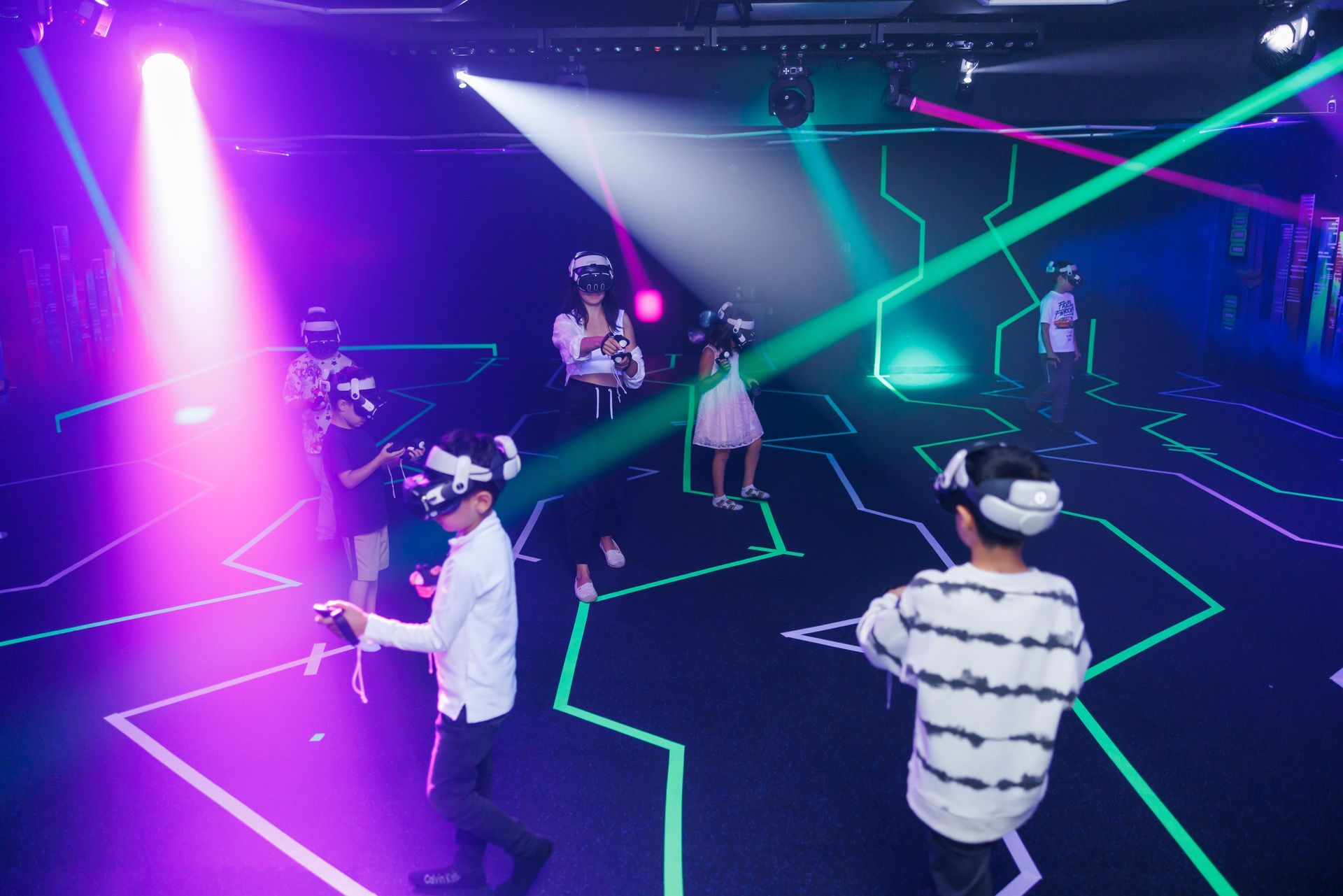People wearing VR headsets navigate an indoor neon-lit gaming space with glowing floor outlines and vibrant spotlights.