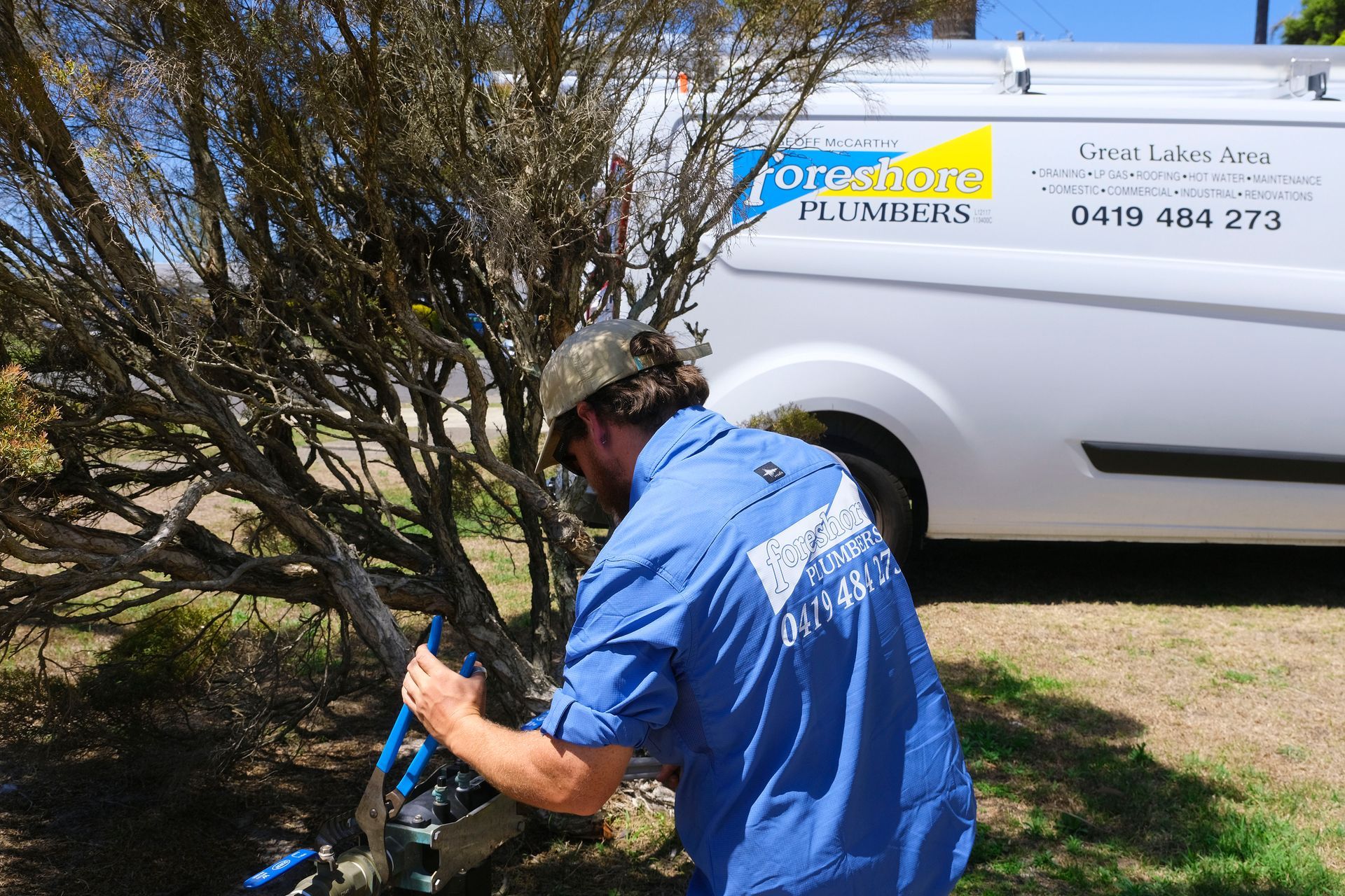 A Man in a Blue Shirt is Working on a Tree in Front of a White Van — Foreshore Plumbers in Tuncurry, NSW