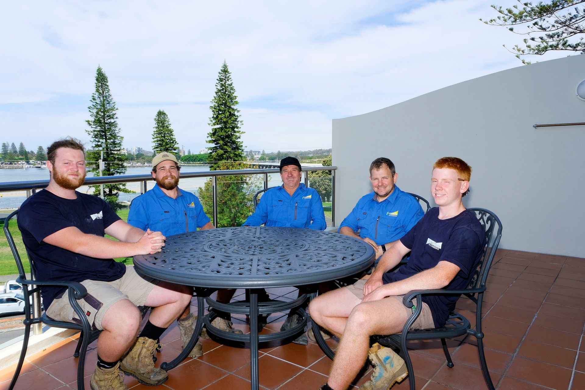 A Group of Men Are Sitting Around a Table on a Balcony — Foreshore Plumbers In Forster, NSW