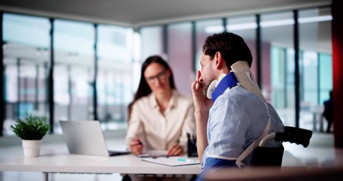 Man in neck brace and wheelchair at office table with woman, documents, and laptop.