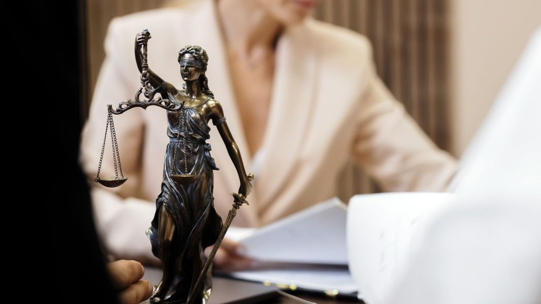 Statue of Lady Justice on a desk, woman in blazer in background, reviewing documents.