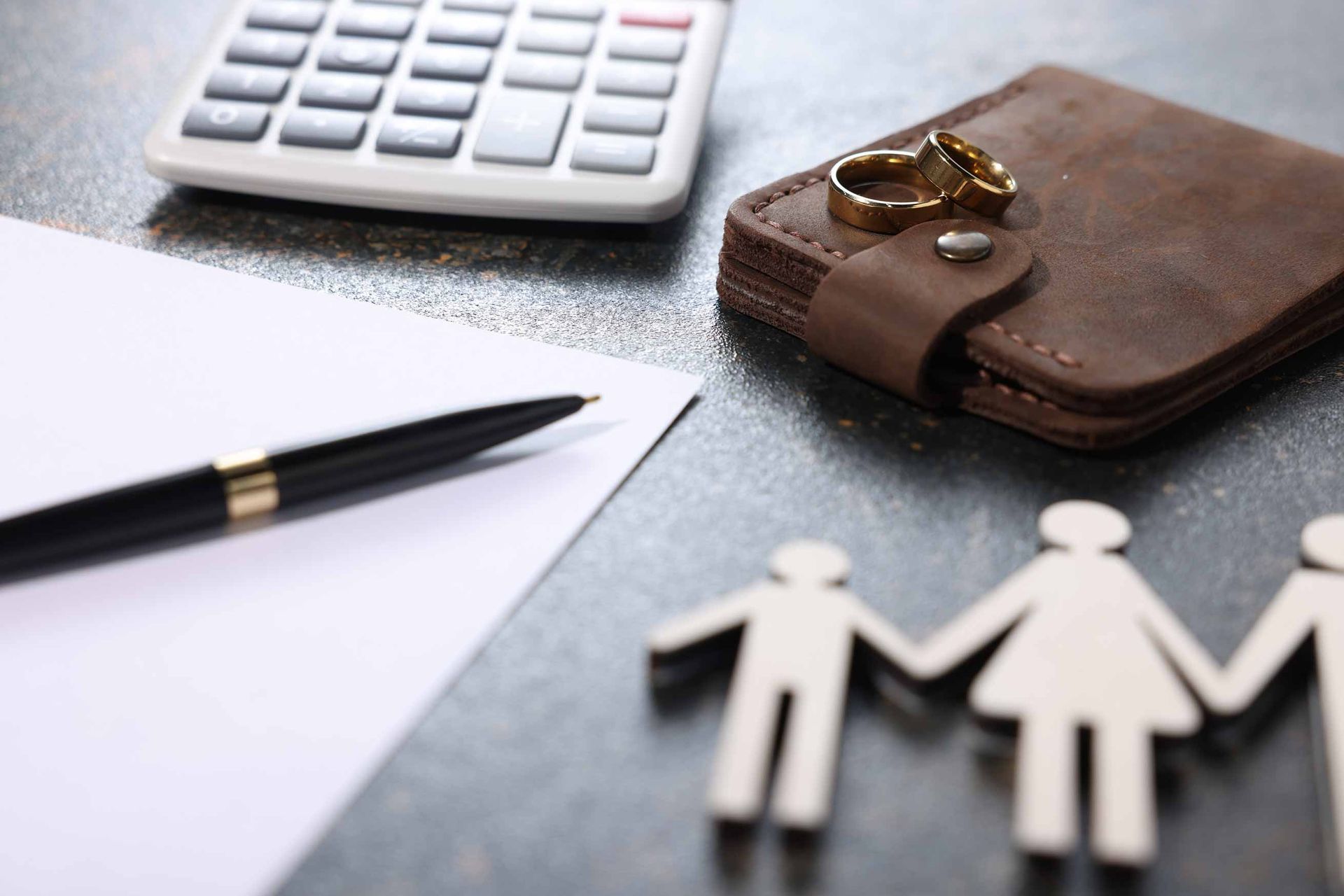 A calculator, a pen on a blank sheet of paper, a leather wallet with two gold wedding rings on top.