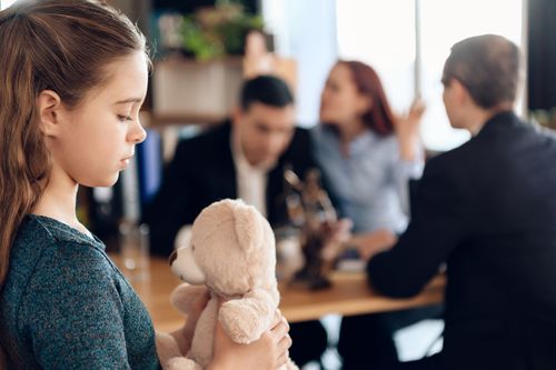 Girl holding a teddy bear looking down, parents arguing in the background at a table.