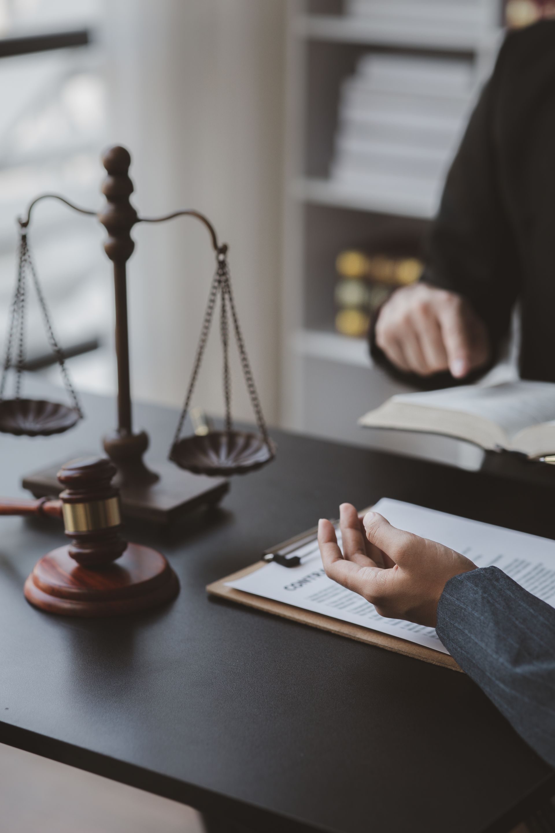 Scales of justice, gavel, and hand on a document at a desk with a blurred figure in a judicial robe.