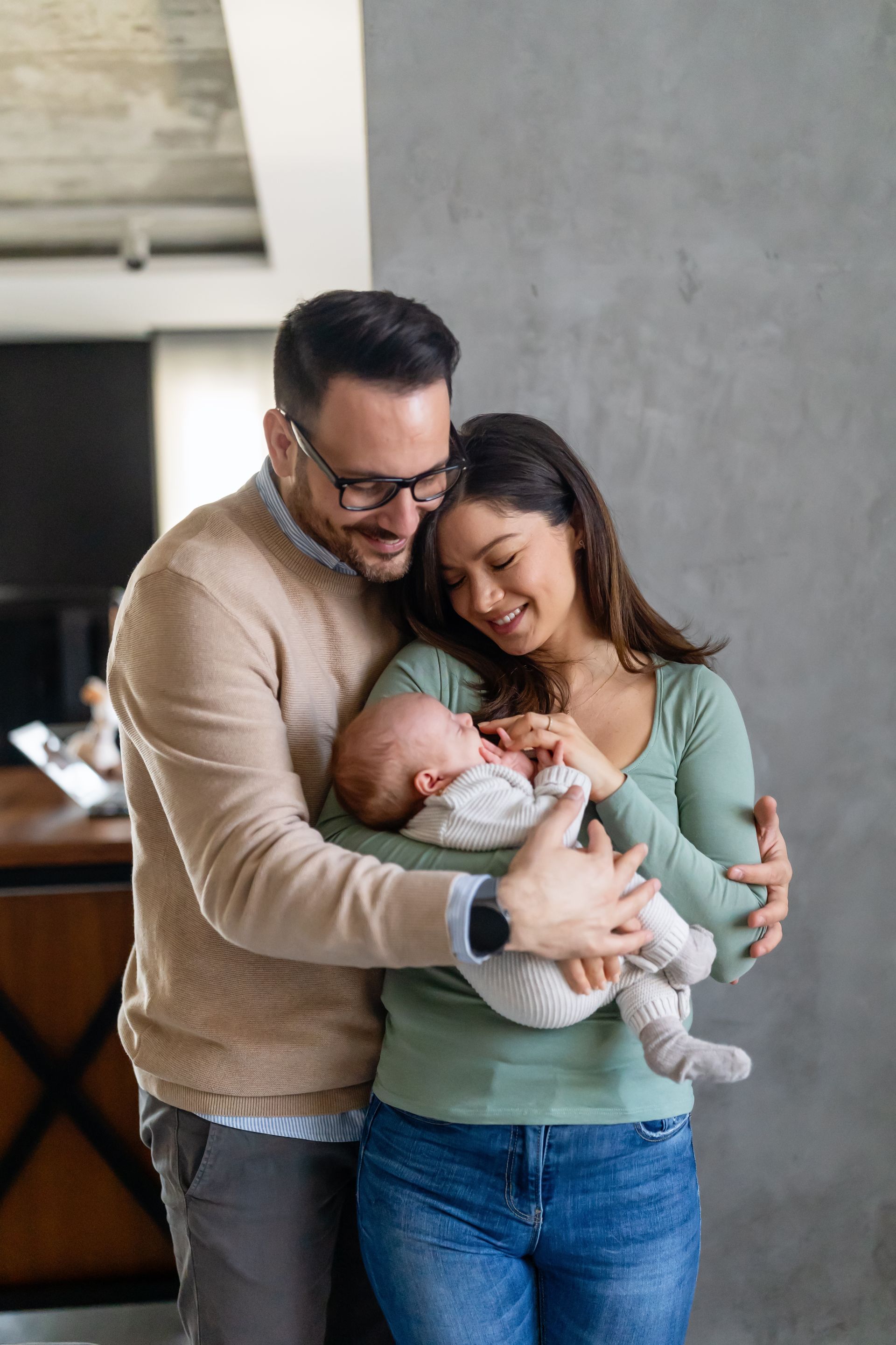 Couple smiling, holding baby. Man in glasses, tan sweater. Woman in green shirt, jeans. Gray wall background.
