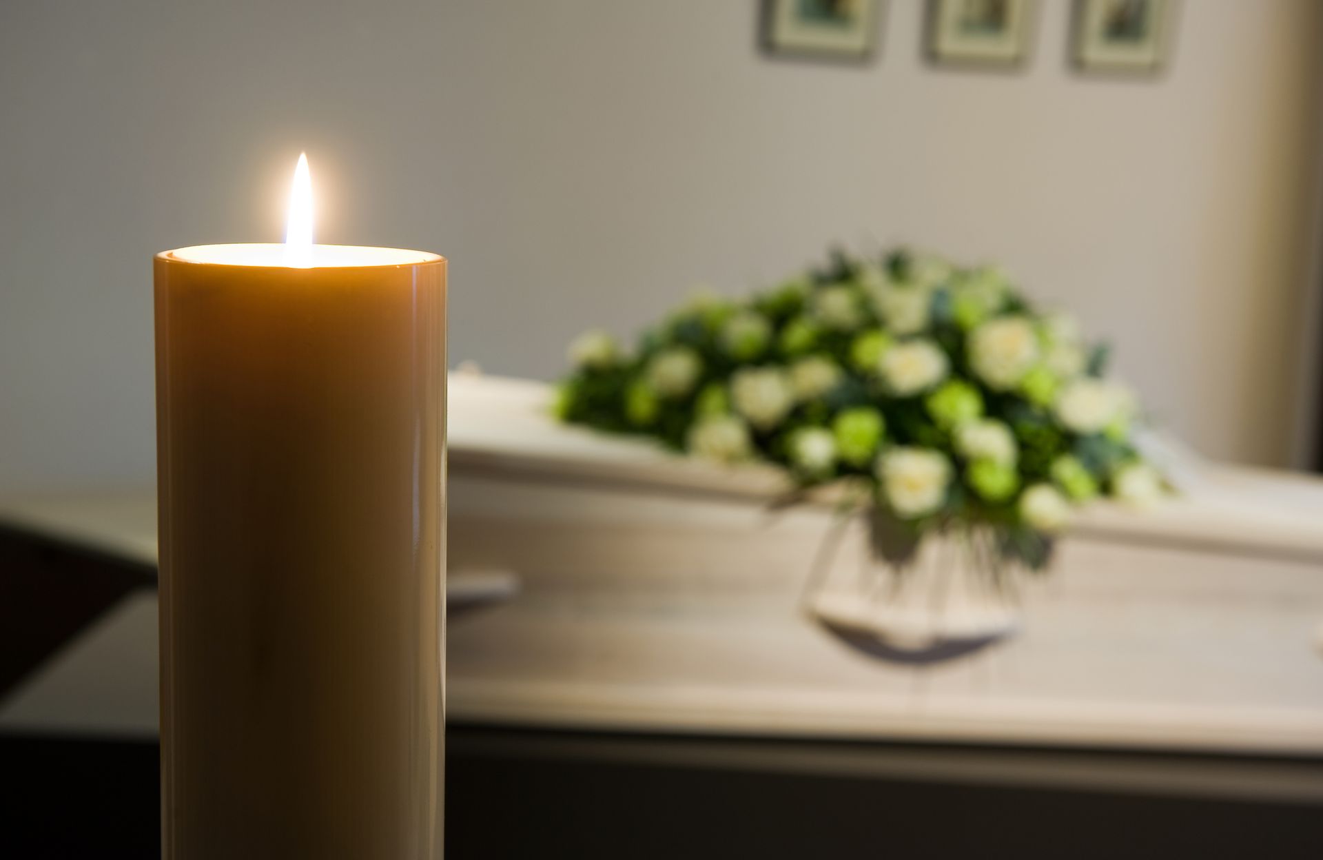 Candle burning brightly in front of a closed white coffin with floral arrangement, soft lighting.