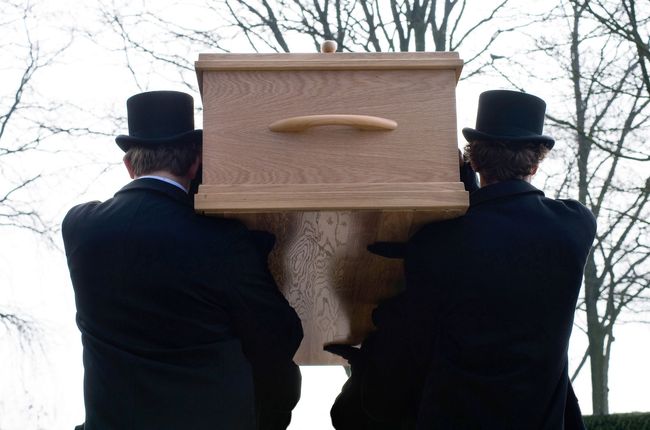 Two pallbearers in black coats and top hats carrying a light-colored coffin outdoors.