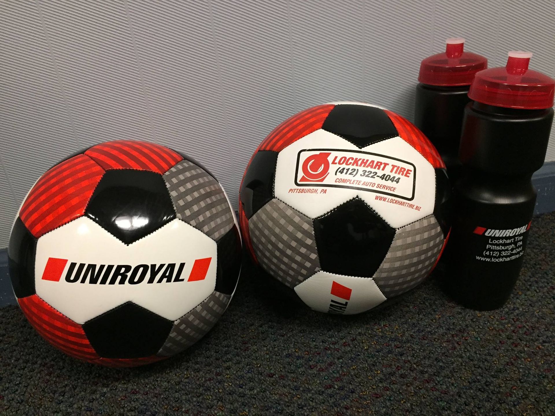 Two black-and-white Uniroyal soccer balls beside two black and red bottles on a textured surface.