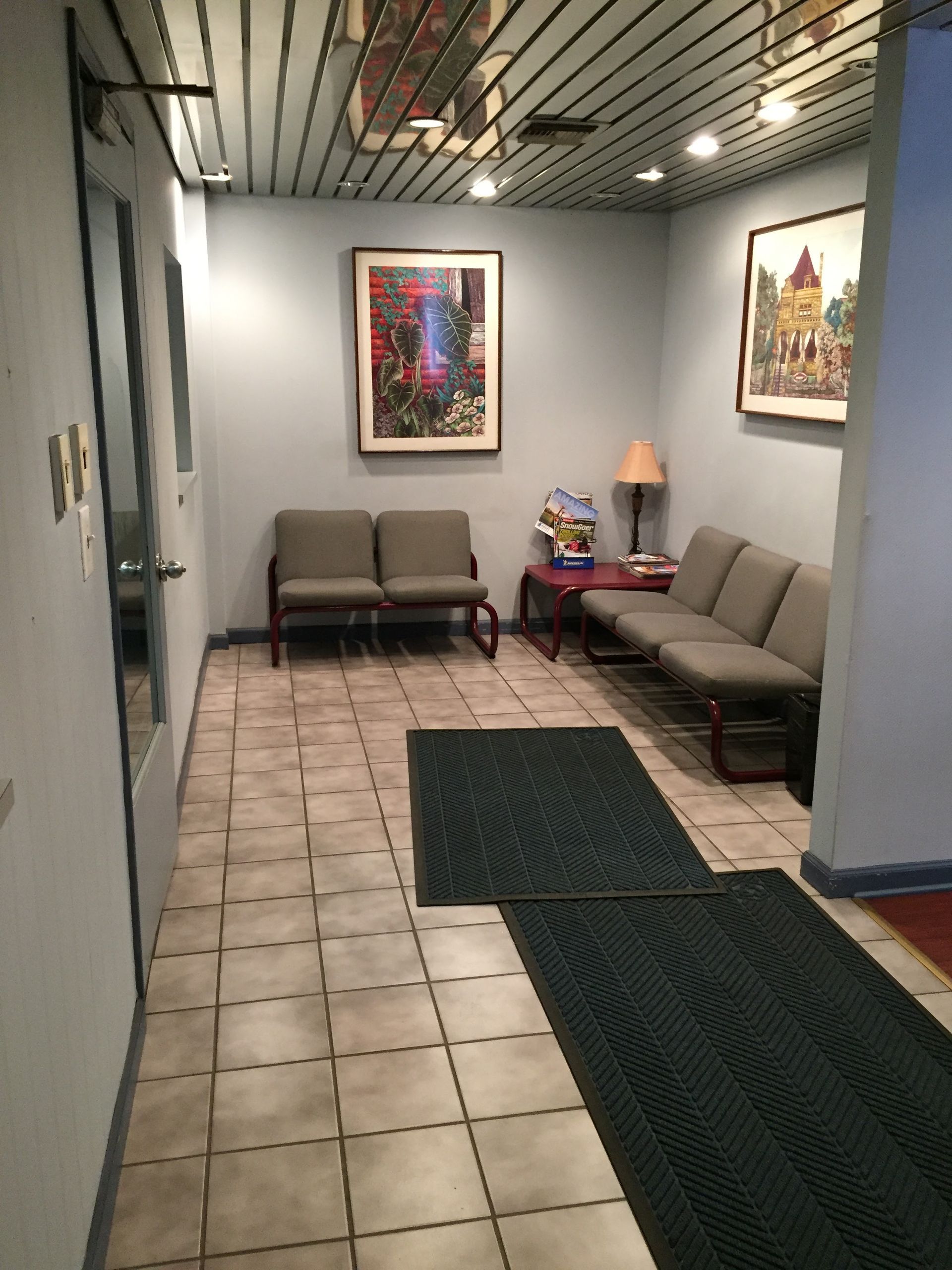 Waiting area with gray chairs, framed art, and dark green floor mats in a tiled hallway.