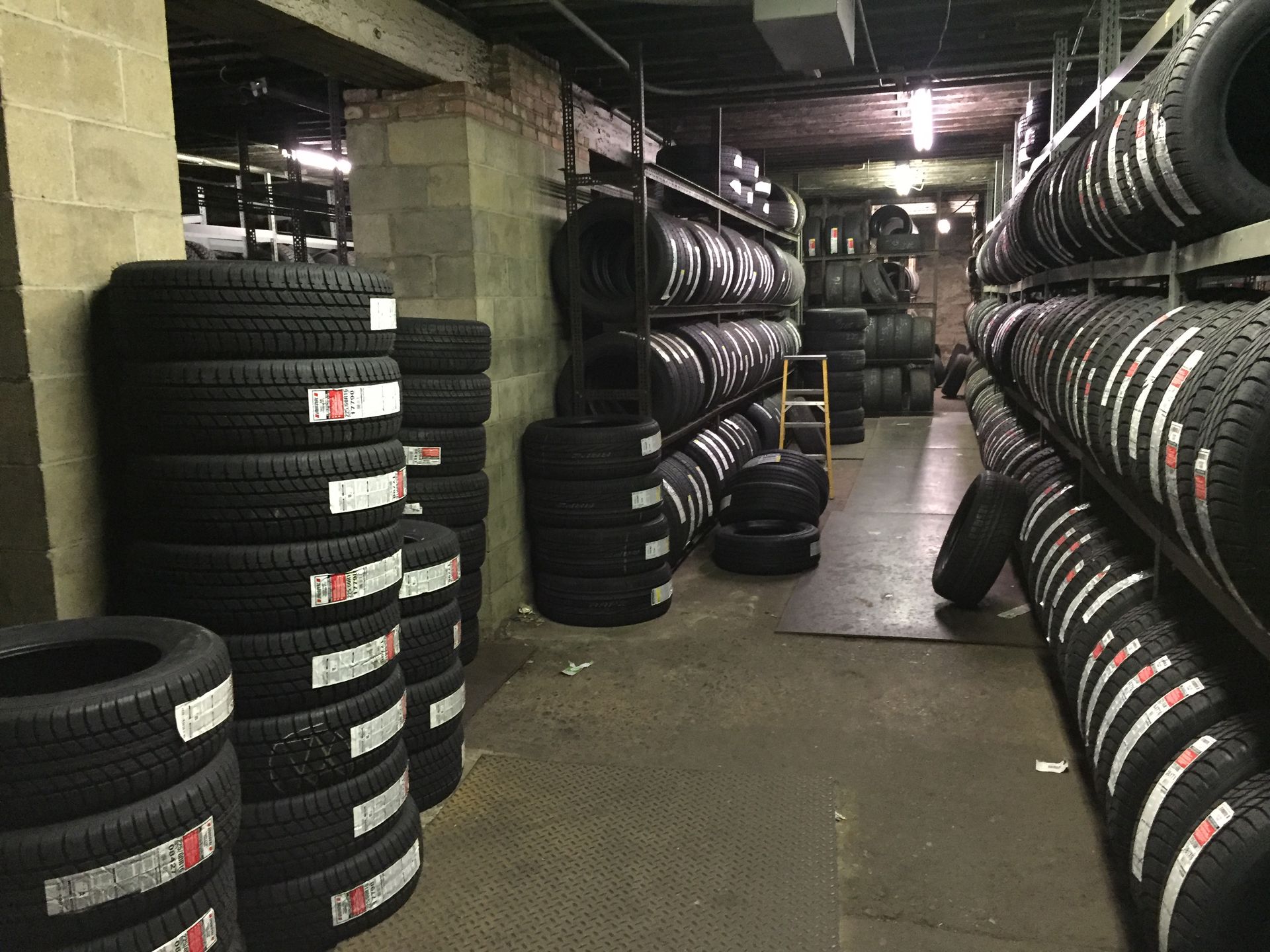 Stacks of black tires lining a dim warehouse aisle, with more tires stored on racks along both sides.