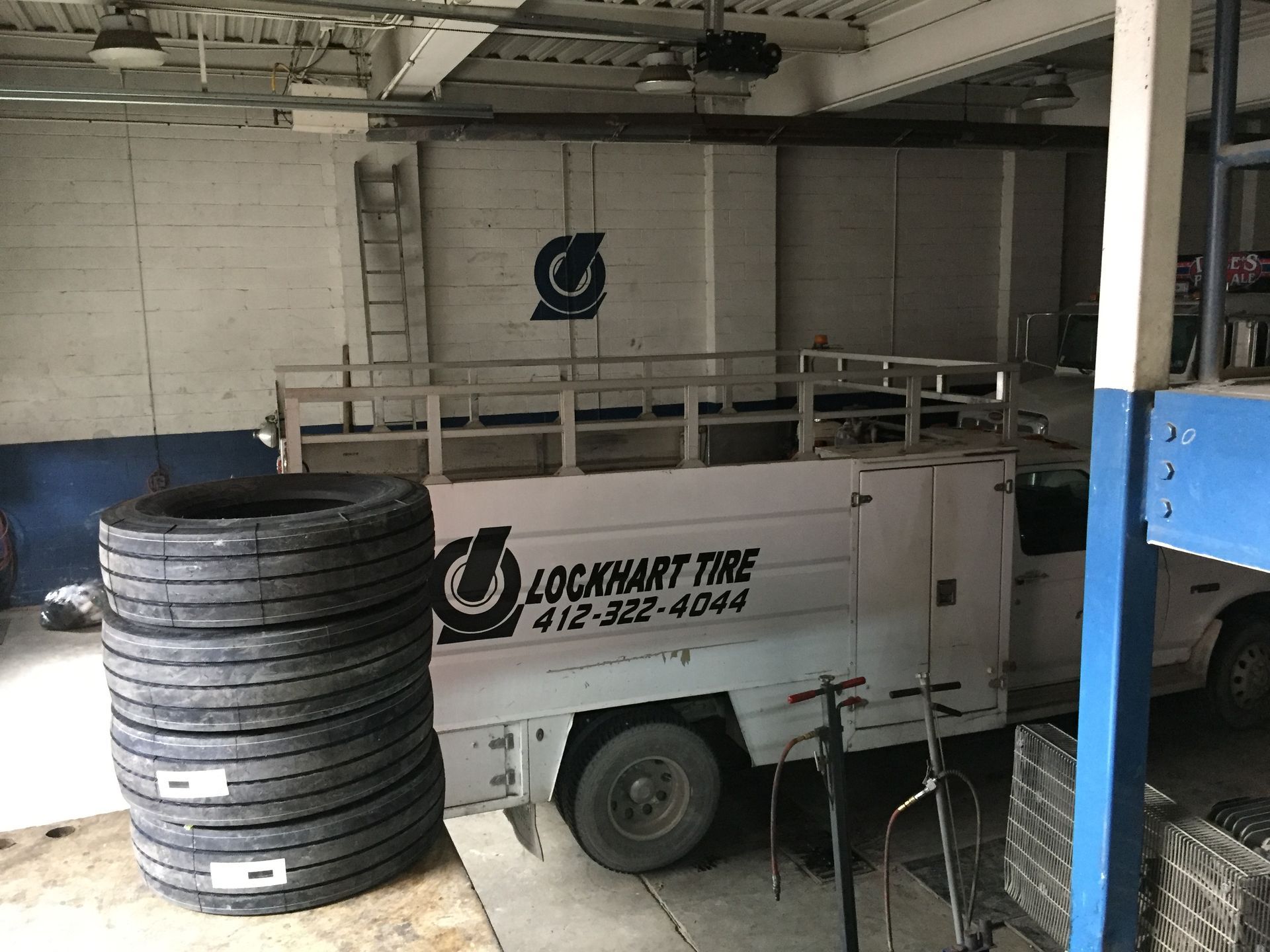 White service truck parked indoors beside stacked tires in a garage, with a blue vehicle partly visible.