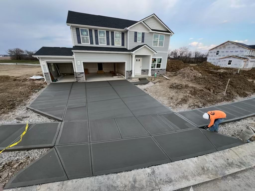 Worker smoothing a large gray concrete driveway in front of a suburban house under construction.