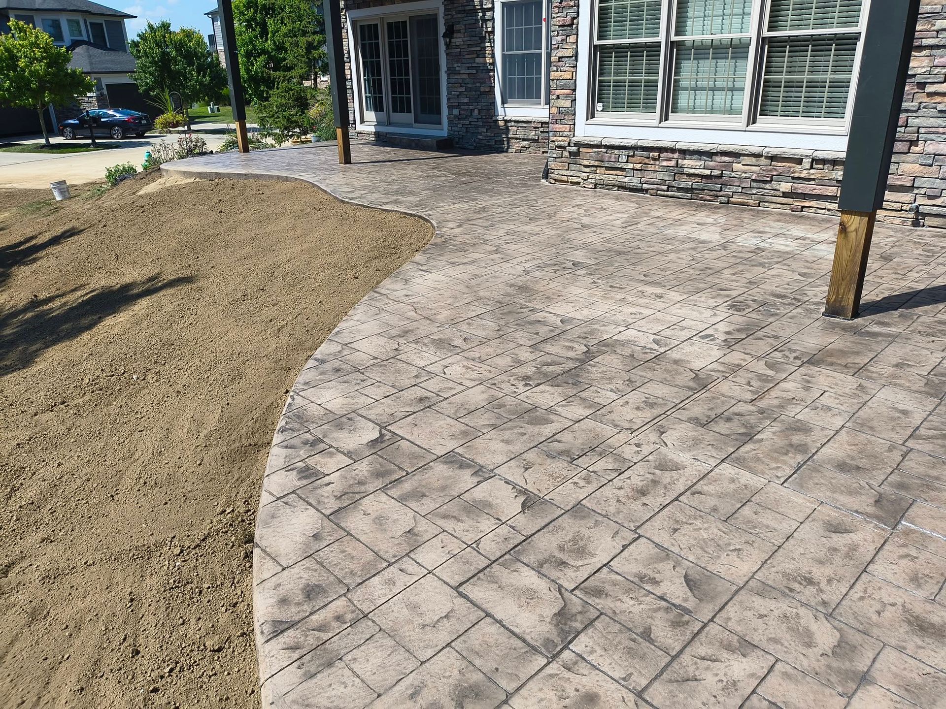 Curved stone patio beside a house, next to a bare dirt landscaping area.