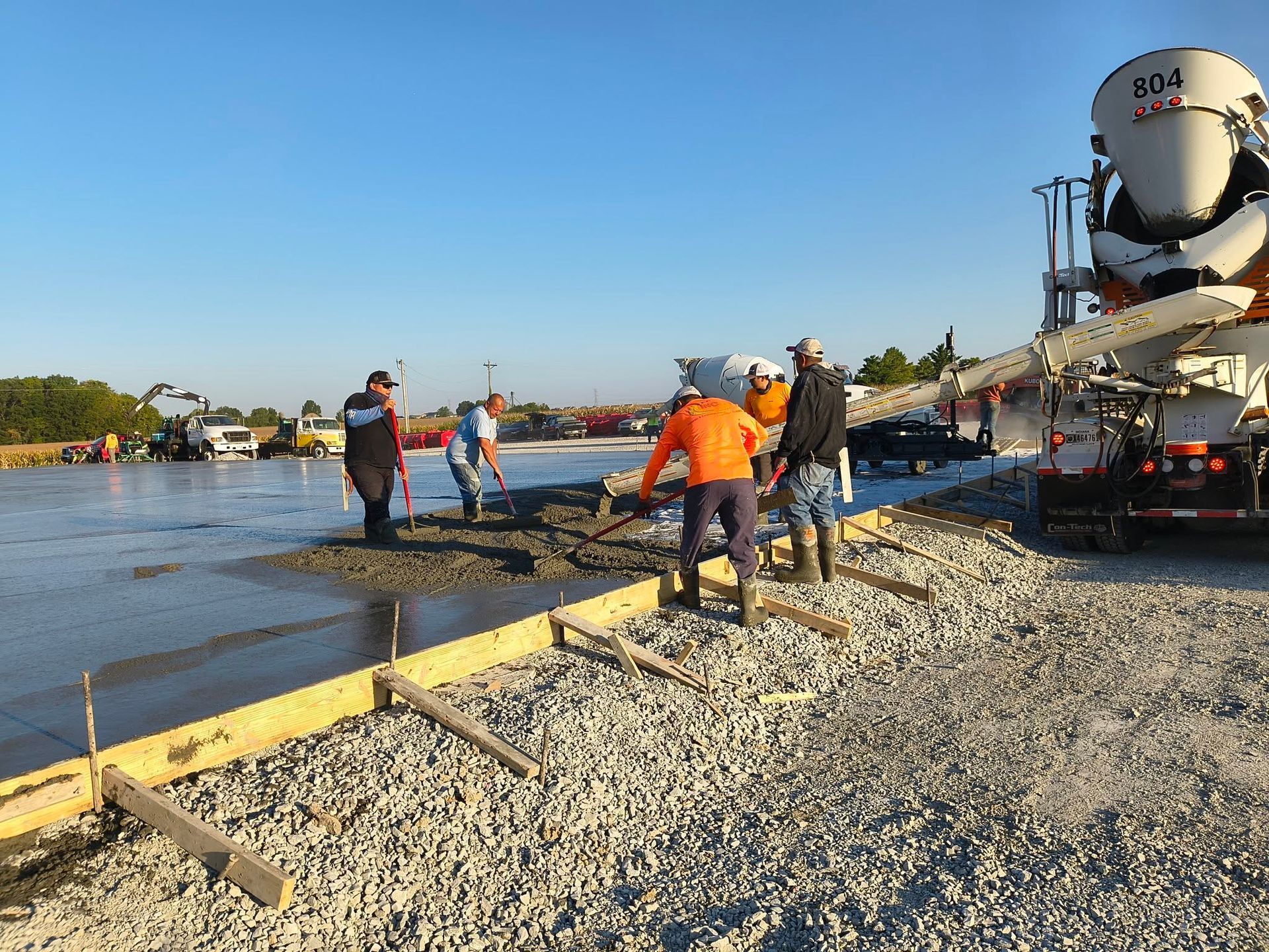 Workers standing by a concrete pour at a construction site beside water, with a cement mixer truck nearby.