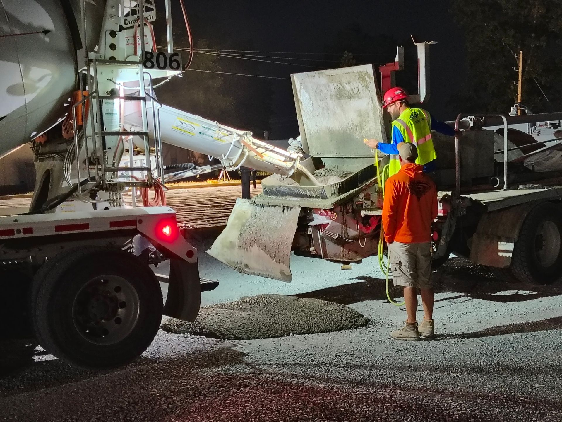 Construction worker inspecting concrete pump truck at night on a gravel road, with debris and lights visible
