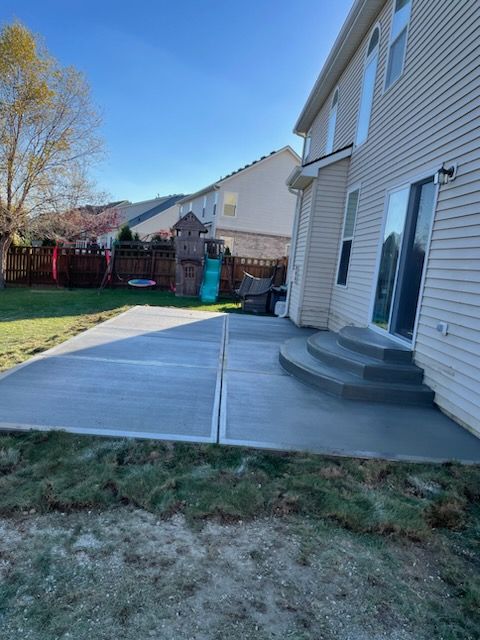 Backyard concrete patio beside a house, with steps, fence, and grass in sunlight