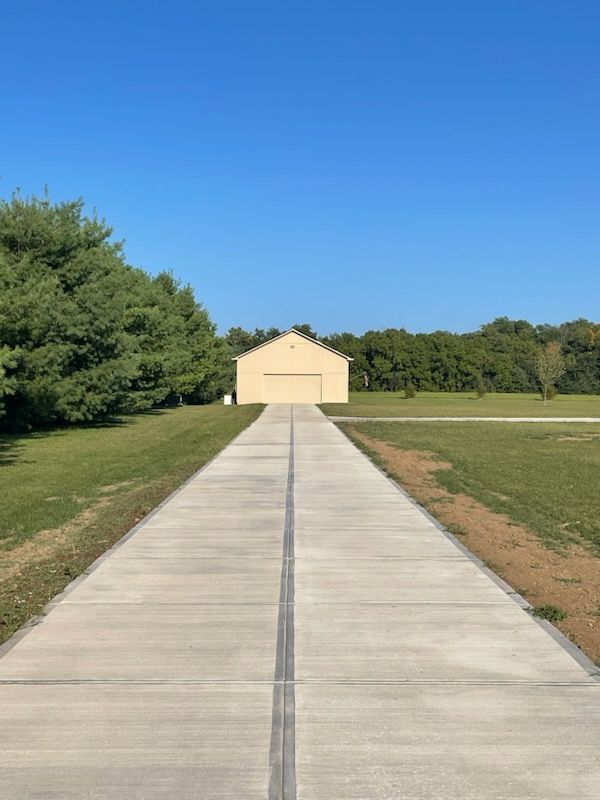Concrete driveway leading to a small beige building under a clear blue sky, lined by green trees and grass.