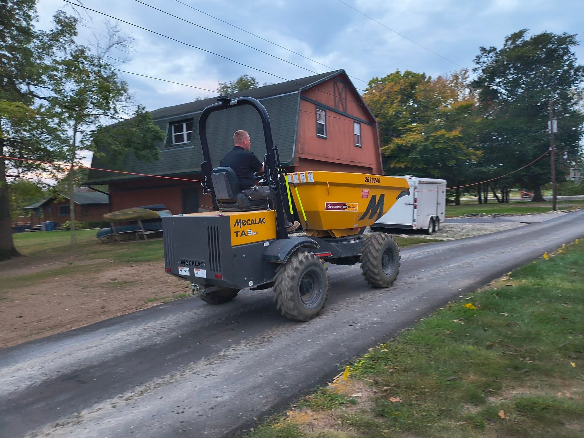 Yellow dump truck on a rural road beside a house and trailer, with a driver visible in the cab.