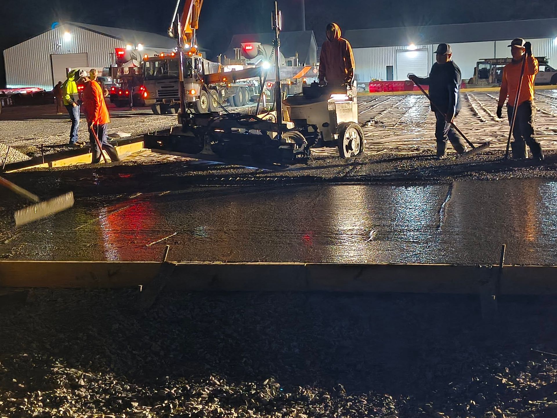 Nighttime roadwork scene with workers in orange safety gear and construction vehicles on a wet street