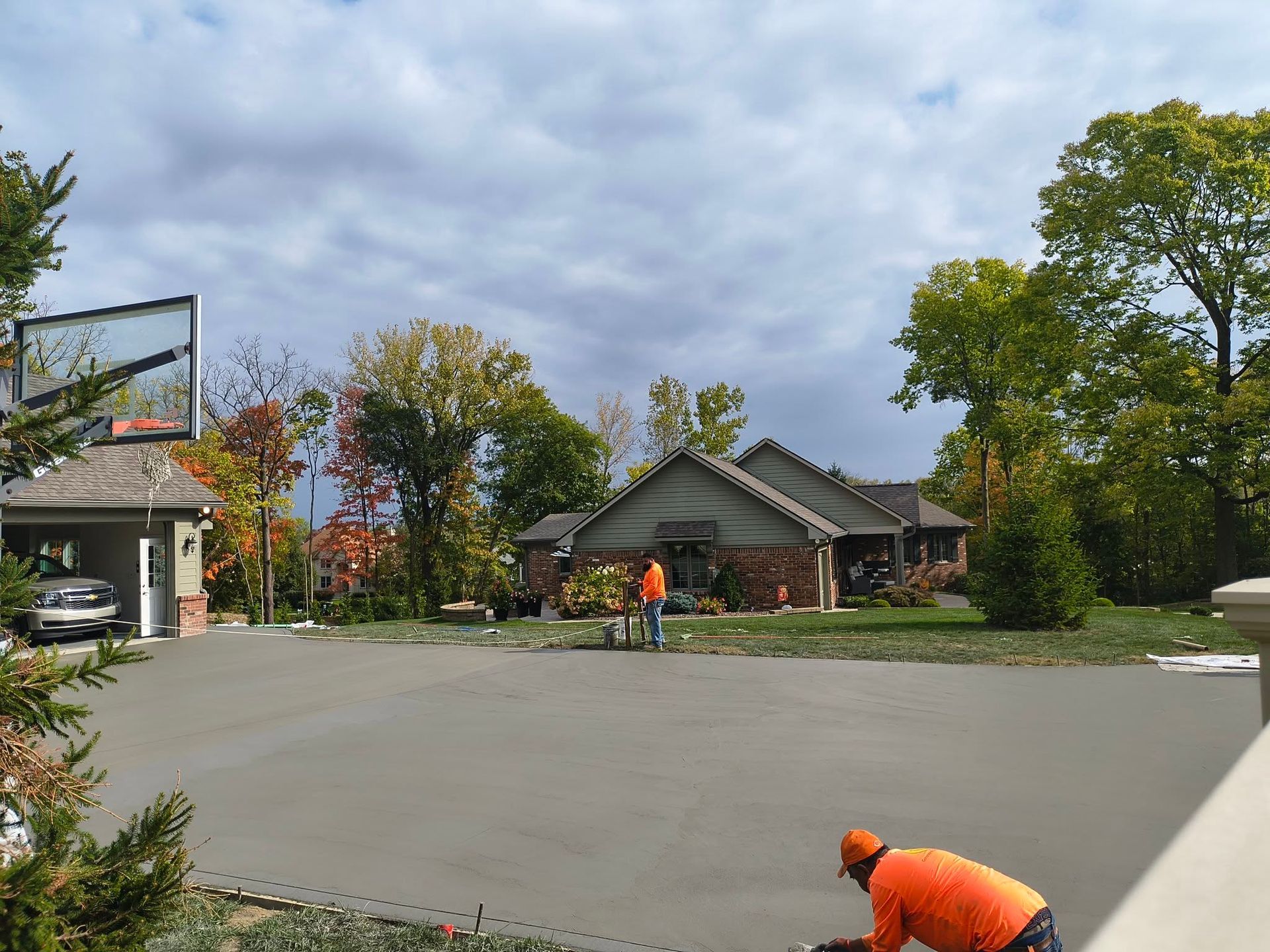 Two workers in orange shirts paving a wide driveway beside a house and trees