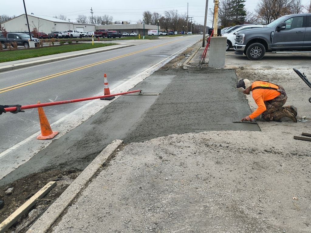 Roadwork crew member in orange kneels beside a torn-up roadside with cones and traffic barriers nearby.