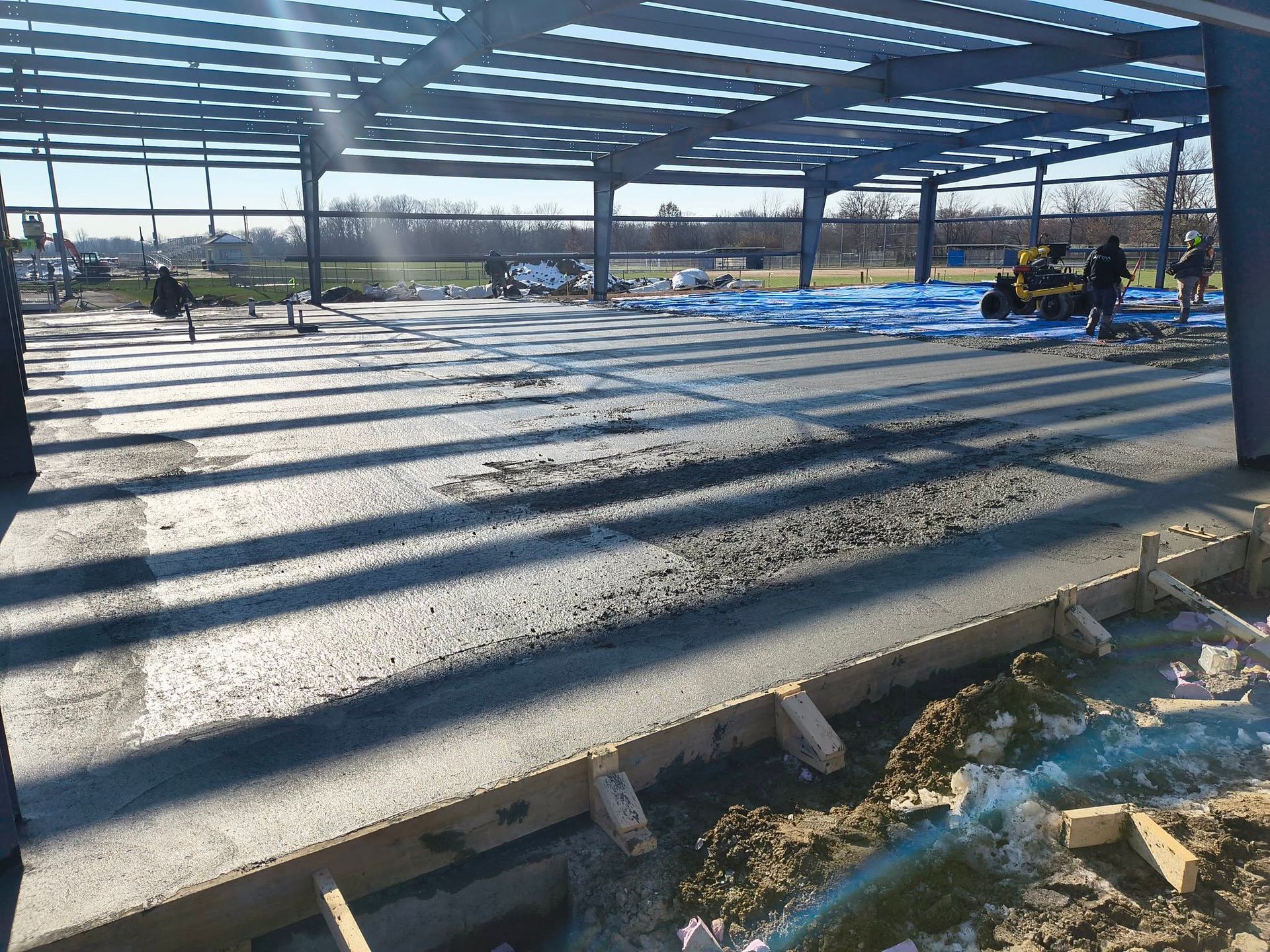 Open-air construction site with concrete floor slab, steel columns, workers, and blue tarps in the background