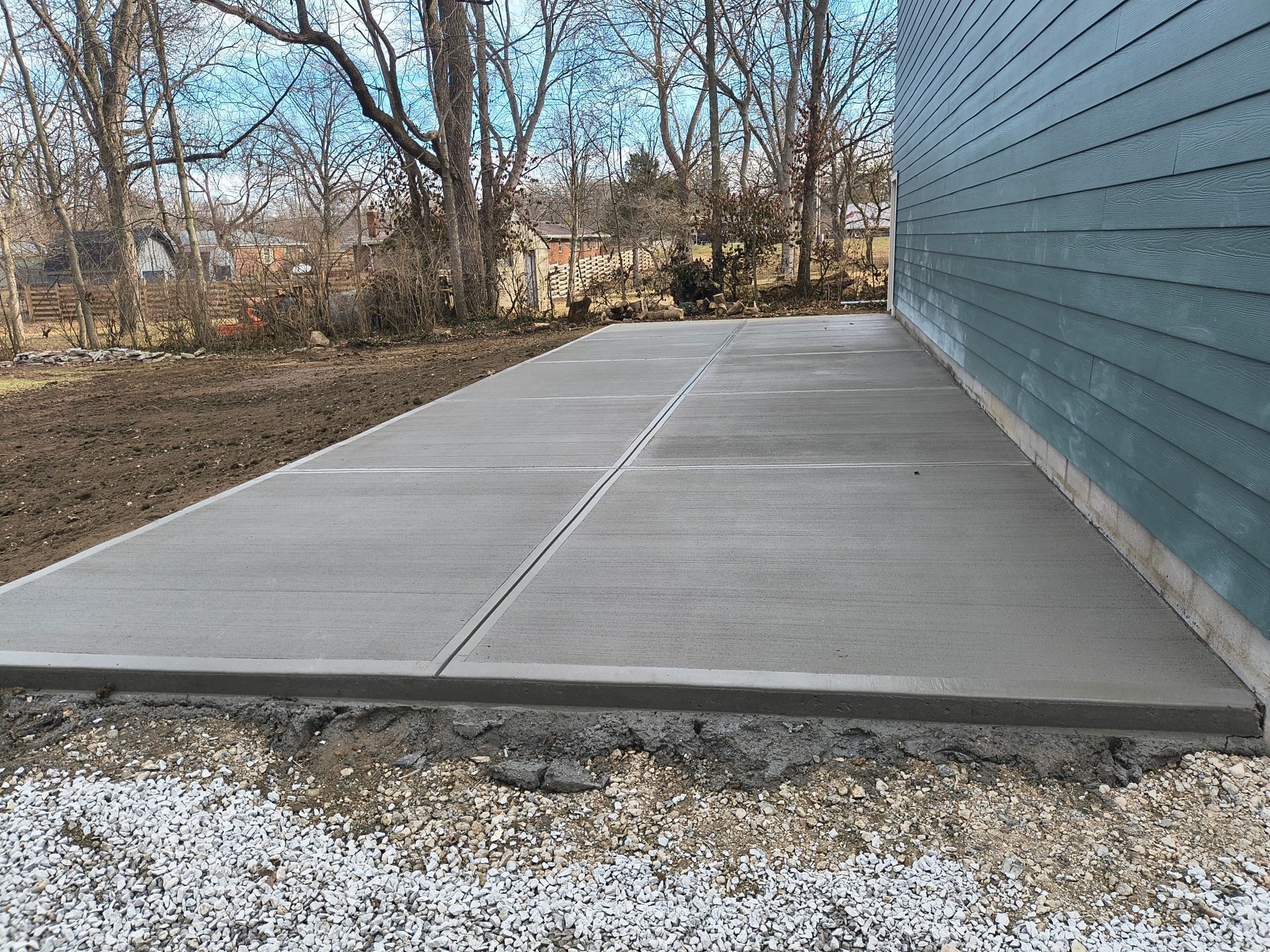 Concrete sidewalk beside a blue house, bordered by bare trees and patchy snow.
