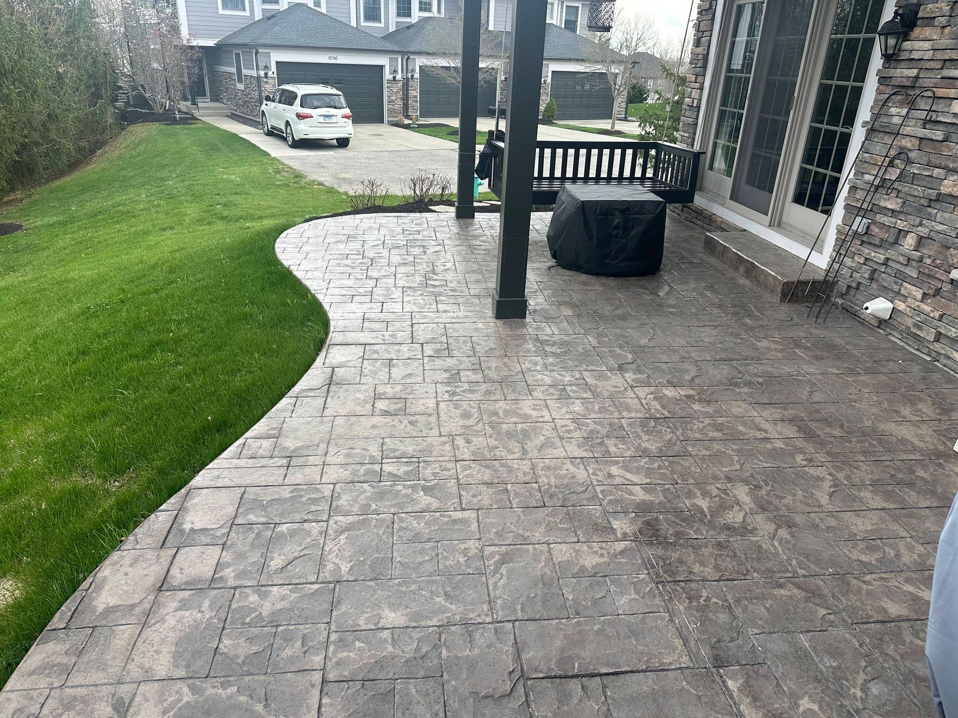 Stone backyard patio beside a house, with green lawn, railing, and driveway visible in the background