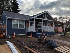 A blue single-story house with a white-railed porch under construction, surrounded by dirt and construction materials.