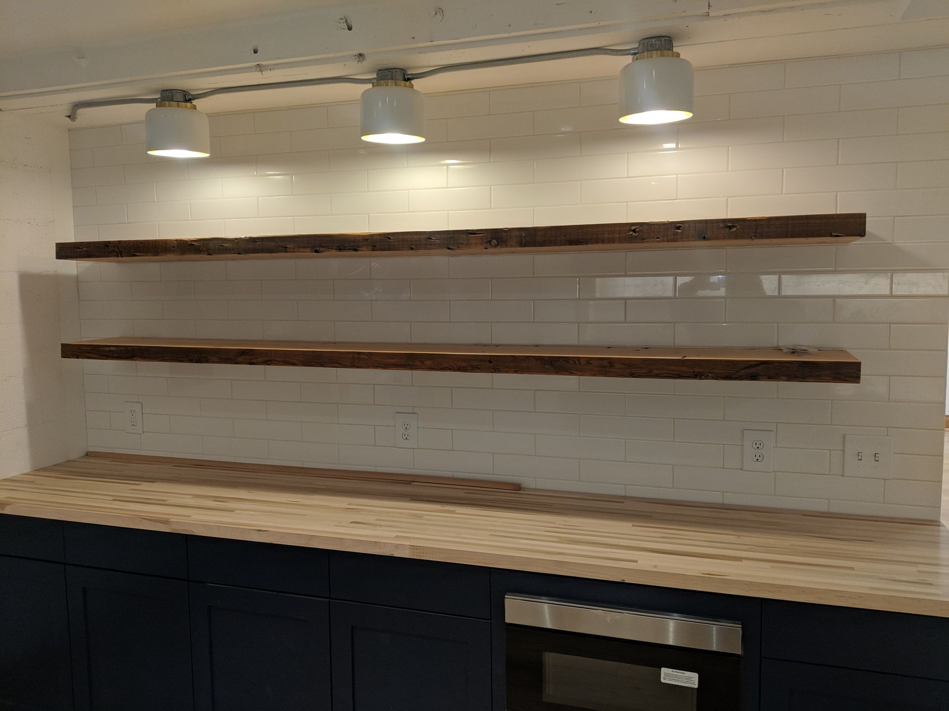 Modern kitchen with white tile backsplash, two wooden shelves, pendant lights, and dark cabinets