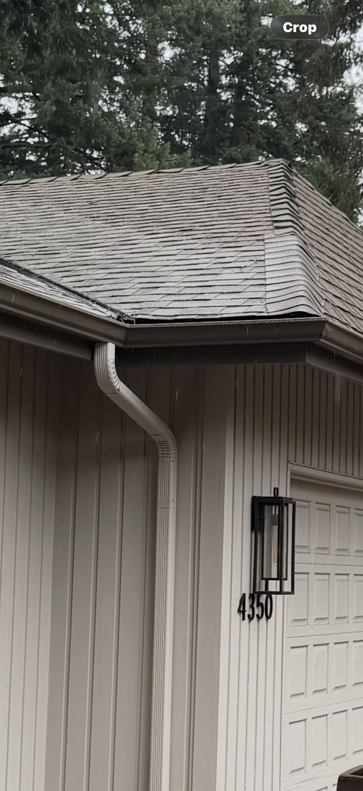 A side view of a house garage, featuring beige vertical siding, a house number, an outdoor light, and worn roof shingles.