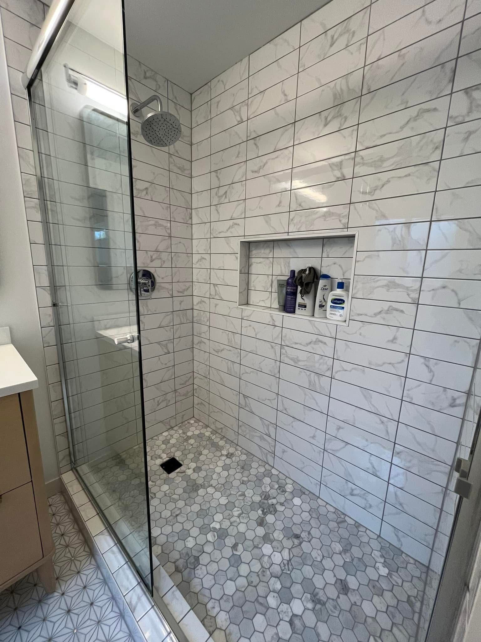 A walk-in shower with white marble-patterned wall tiles, a built-in storage niche, a glass door, and pebble floor tiling.