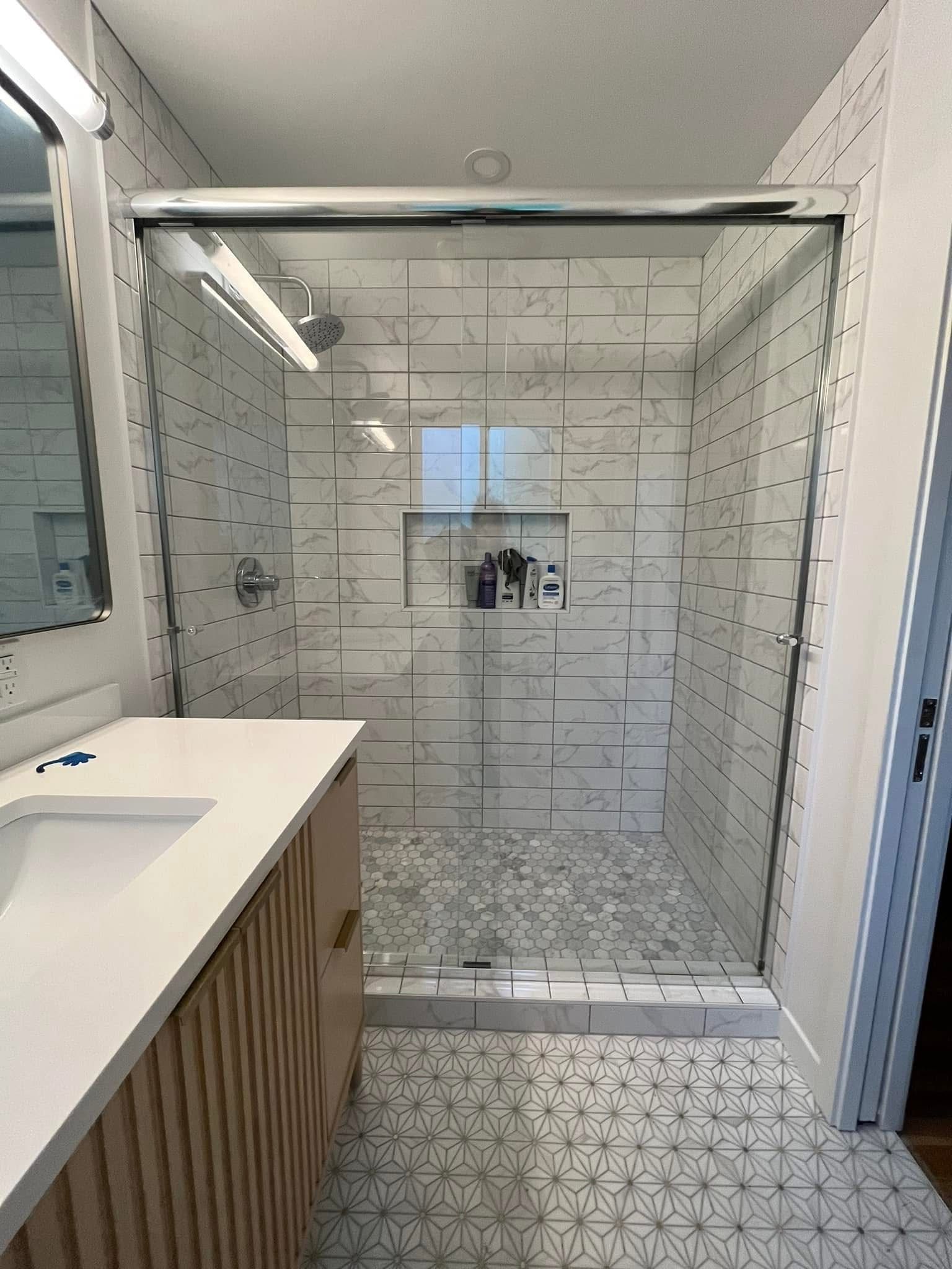A modern bathroom featuring a glass-enclosed shower with white marble-patterned tile, a tiled floor, and a wood vanity.