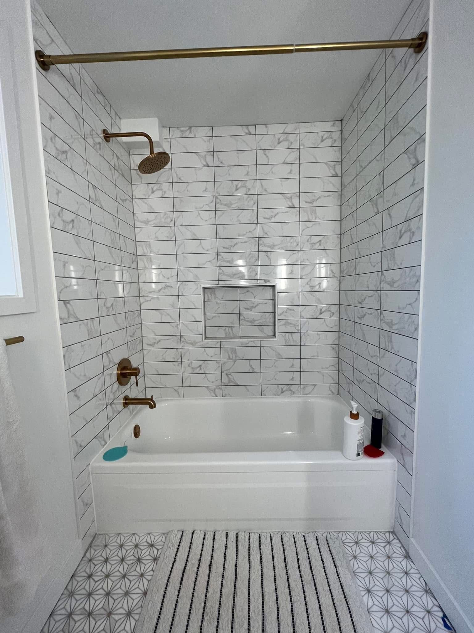 A bathtub and shower area with white patterned wall tiles, gold fixtures, a recessed shelf, and a patterned floor mat.