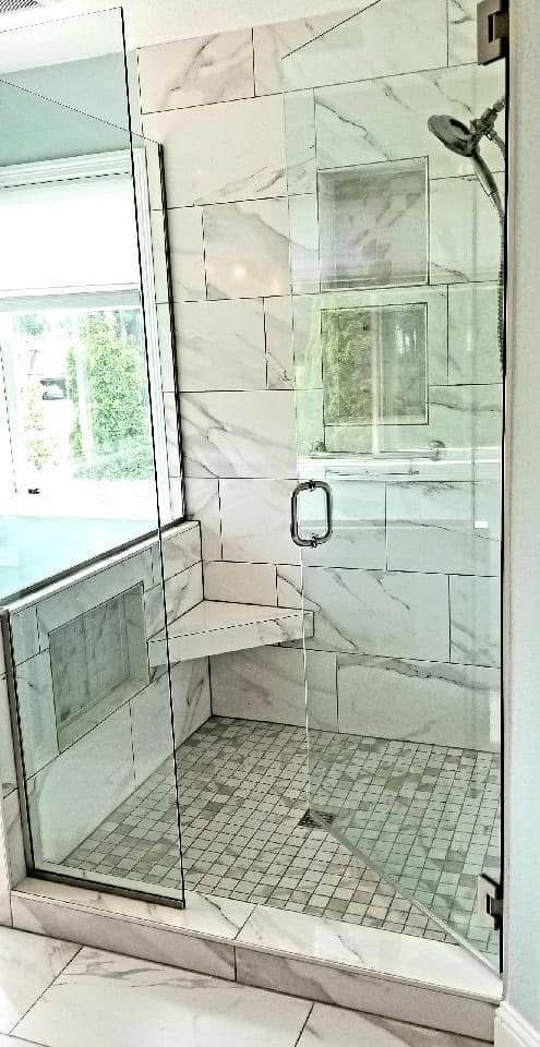 A walk-in shower with marble-look wall tiles, a mosaic tile floor, a built-in corner seat, and a glass door.
